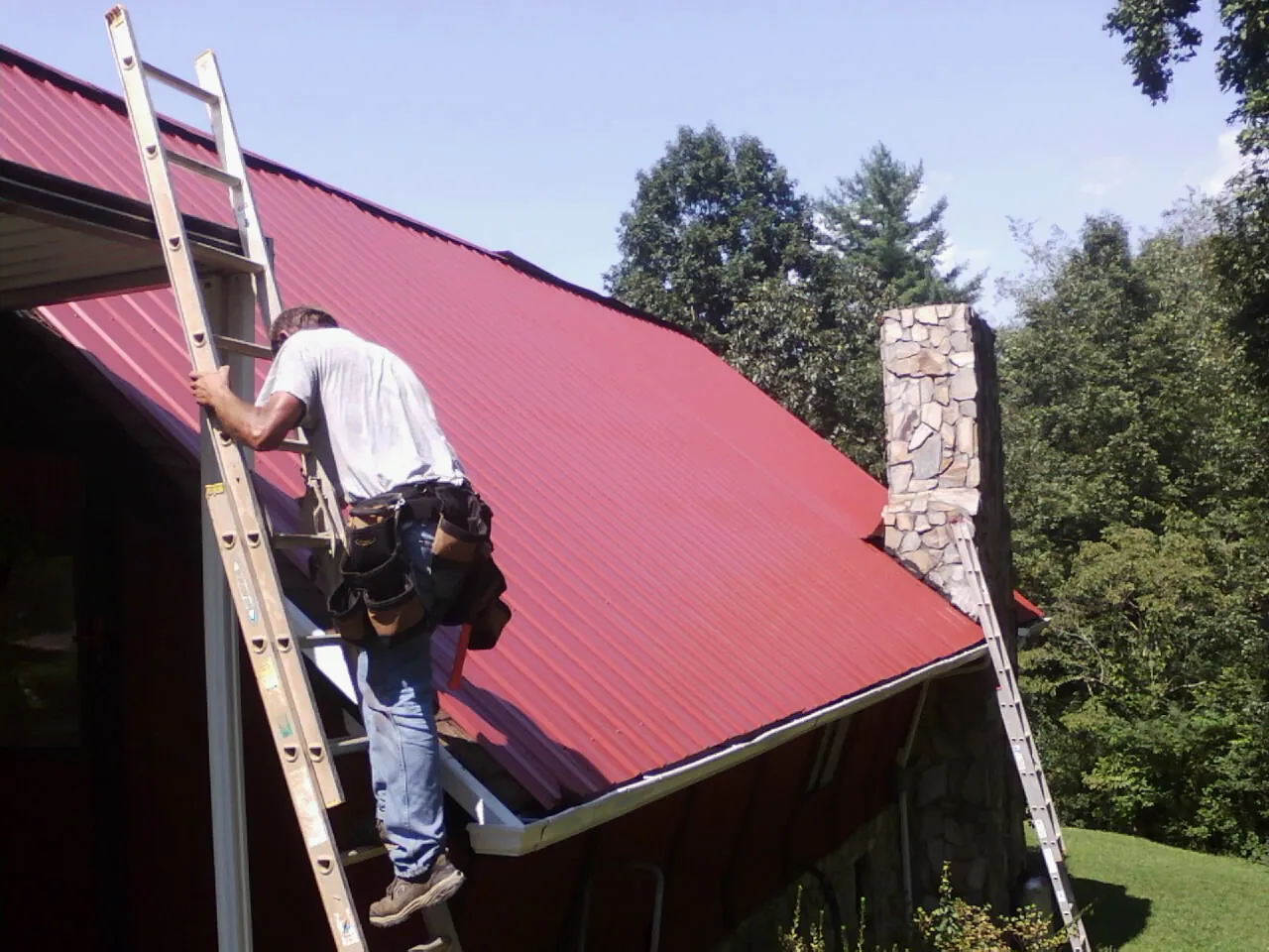 Roofer on a ladder beside a red metal roof and stone chimney — Soffit & Fascia Repair by Roofing Brothers in Broadalbin